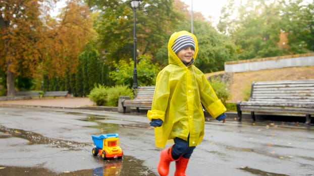 Portrait of smiling cheerful toddler boy running over puddle at park with his toy truck.