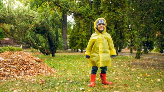 Happy smiling little boy wearing yellow raincoat and red rubber boots after rain at park.
