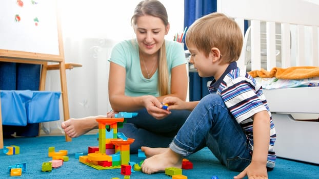 Portrait of happy smiling boy with young mother playing on floor with colorful toys.