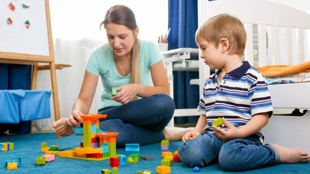 Young mother helping her little son assembling toy constructor on carpet at playroom.