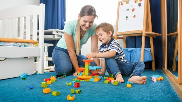 Happy smiling mother sitting on floor with her little son and playing with colorful constructro blocks.