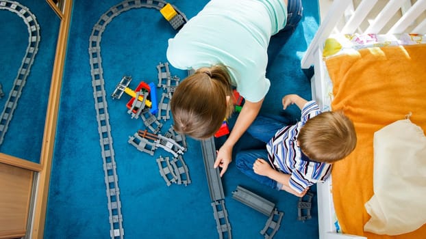 Top view on young mother with little son building toy railroad from colorful bricks and blocks.