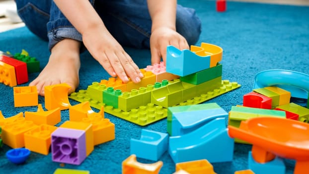 Closeup image of little toddler boy sitting on carpet at home and playing with colorful constructor blocks.