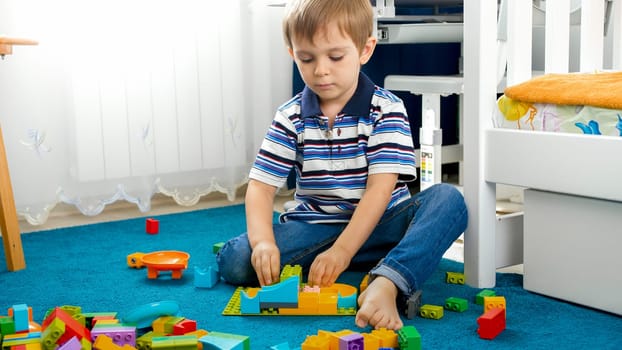 Portrait of little toddler boy sitting on floor in bedroom and playing with toy constructor.