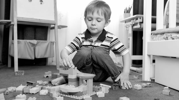Black and white portrait of little toddler boy playing with constructor blocks and bricks on carpet at his room.