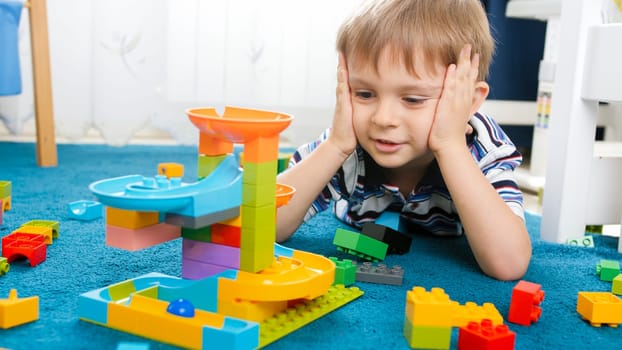 Portrait of cute toddler boy lying on floor and looking on colorful construction from plastic bricks and blocks.