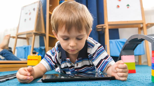 Portrait of little 4 years old boy lying on carpet in his room and using digital tablet computer.