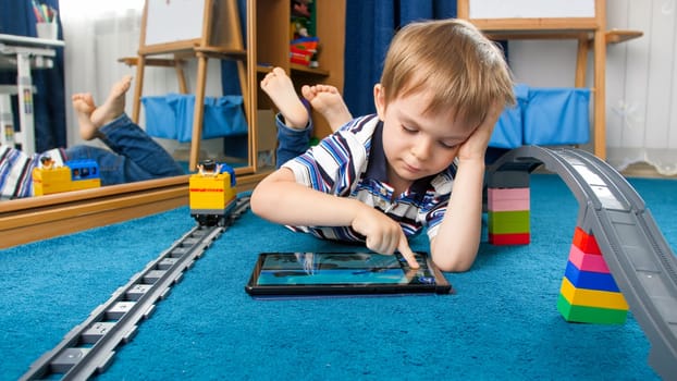 Cute little boy lying on floor and playing educational games on tablet computer.