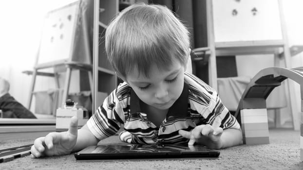 Portrait of cute little boy playing games on tablet lying on carpet at living room.
