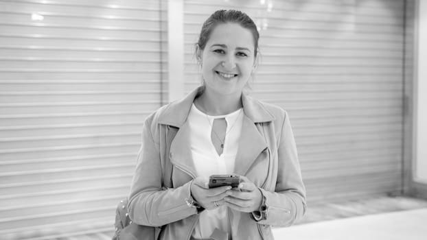 Black and white portrait of smiling young woman holding smartphone and looking in camera.