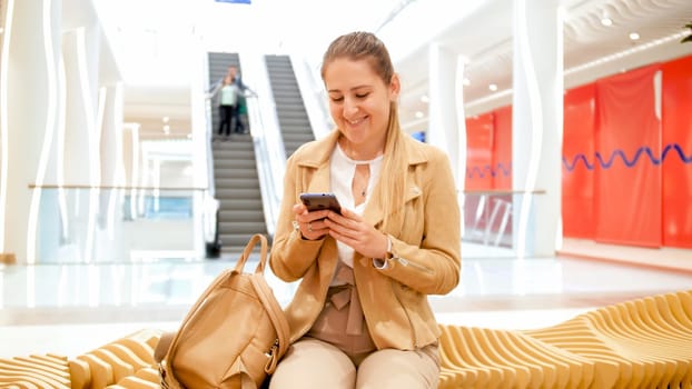 Beautiful smiling young woman sitting on bench in shopping mall and using smartphone.
