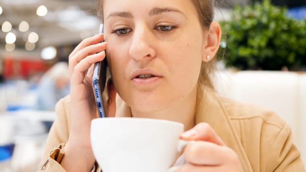 Portrait of busy young woman drinking coffee in cafe and talking by phone.