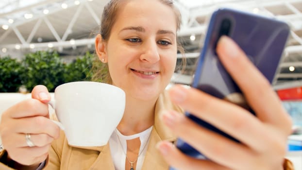 Closeup image of smiling young woman drinking coffee and holding smartphone.