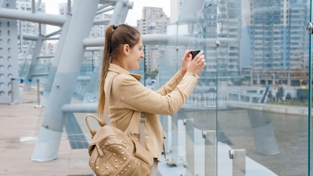 Portrait of beautiful young woman making photograph of river and city skyline on smartphone from glass bridge.
