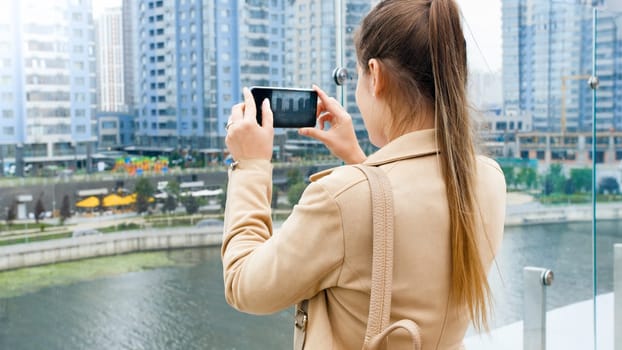Beautiful young woman standing on bridge over the river and making photographs of city skyline on smartphone.