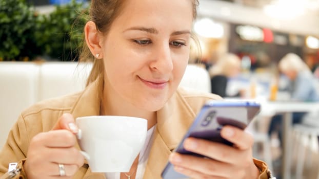 Portrait of young woman drinking coffee and browsing internet on smartphone at cafe.