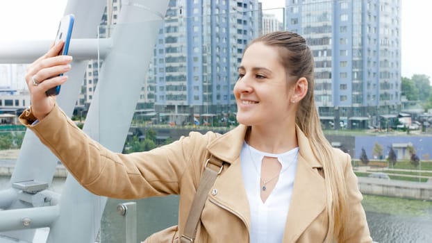 Portrait of beautiful smiling woman taking selfie image on smartphone against the river and city skyline.
