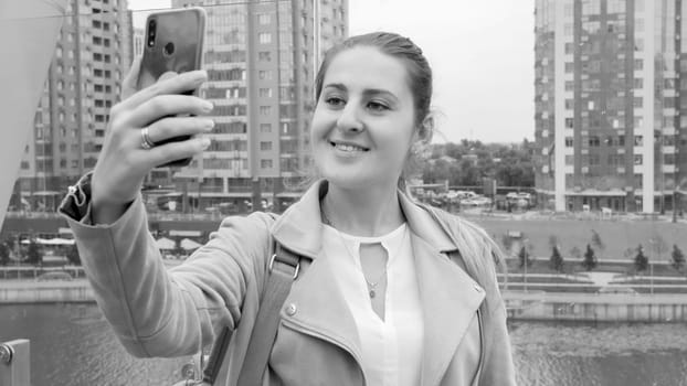 Black and white portrait of beautiful smiling young woman making selfie on her smartphone against modern skyscrapers and river.