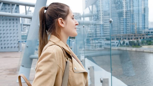 Portrait of beautiful smiling woman standing on bridge and looking on modern buildings and calm river.