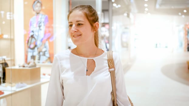 Portrait of smiling young woman choosing new clothes in shopping mall behind glass showcases.
