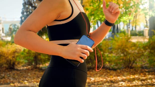 Closeup photo of young woman holding smartphone in hand while running in autumn park and listening music.