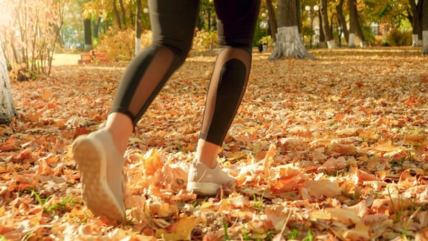 Closeup image of female feet running on fallen yellow tree leaves at autumn park.