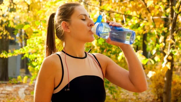 Portrait of beautiful young woman drinking water during sports training in autumn park.