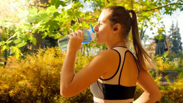 Portrait of beautiful sexy woman with ponytail drinking water from bottle while running and jogging in autumn park.