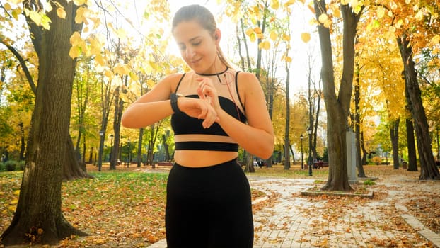 Portrait of smiling sporty woman using smartwatch or fitness tracker during sport exercises in autumn park.