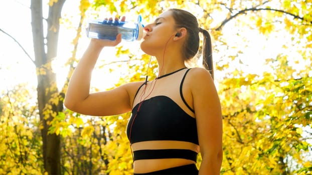 Portrait of beautfiul sporty woman drinking water after fitness training in autumn park.