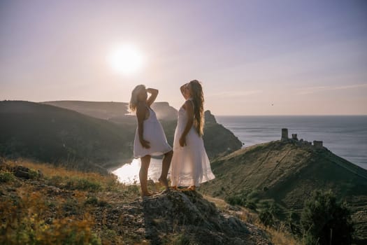 Two young girls are standing on a hillside, one of them wearing a white dress. The sun is shining brightly, creating a warm and inviting atmosphere. The girls seem to be enjoying their time together