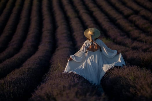 Here's a caption under 150 characters describing the image: A woman in a white dress and wide-brimmed hat walks through a vast lavender field at sunset. The rows of purple flowers stretch to the horizon.
