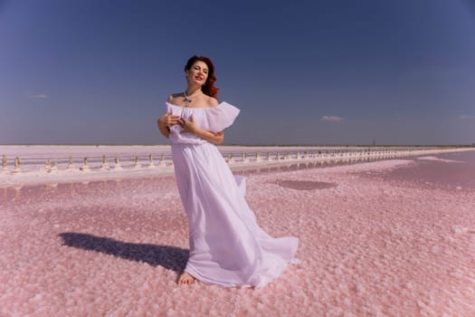 A woman in a white dress stands on a pink salt flat, her hair flowing in the wind, with a clear blue sky above.