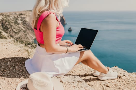 A woman is sitting on a rock by the water with a laptop open in front of her. She is typing on the laptop and she is focused on her work. Concept of productivity and concentration