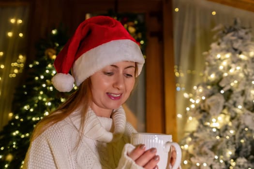 A woman in a Santa hat smiles, holding a white mug in front of a decorated Christmas tree.