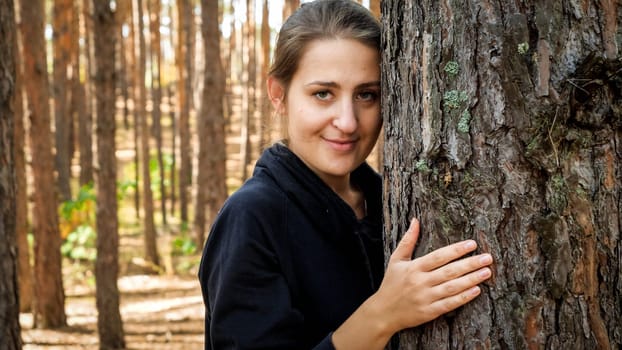 Portrait of smiling young woman huging pine tree in forest and looking in camera. Concept of love, ecology protection and harmony with nature.