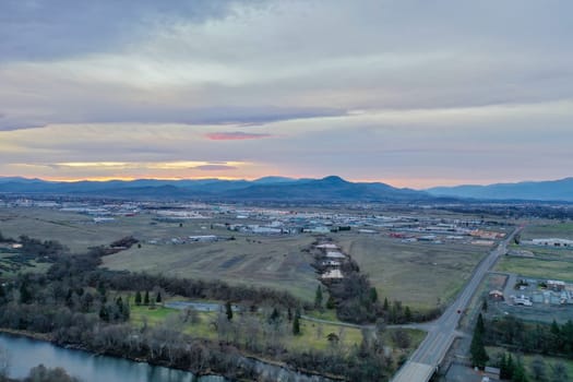 Peaceful wide fields surrounded by trees with mountains as background under cloudy sky. Aerial view of cloudy skyline above trees and roads. Rural town landscape