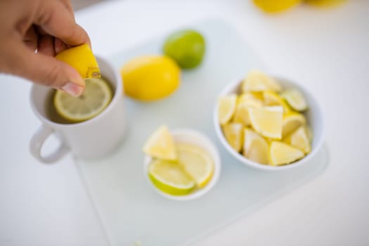Female hand squeezing a lemon into cup of lemon tea next to lemons and limes slices. White mug and citric fruit on white table. Sweet citric beverages