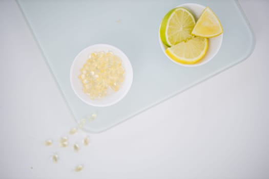 Top view of bowl of transparent vitamins next to bowl of lemon and lime slices on white table. Capsules and chopped citric fruit above transparent board. Vitamin C nutrition