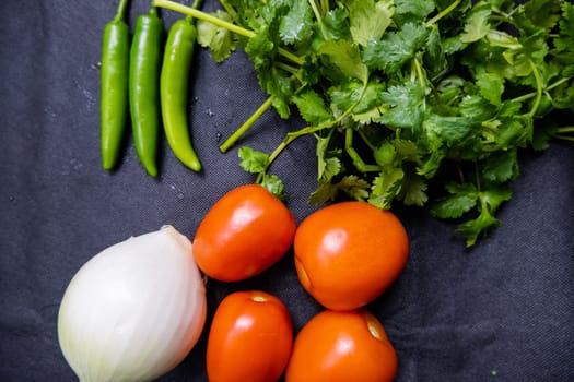 Top view of three green chili peppers, onion, four tomatoes, and coriander on table. Fresh and aromatic vegetables and herbs on dark blue surface. Healthy meal preparation