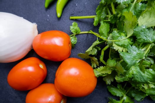 Top view of four tomatoes, green chili peppers, onion, and coriander on table. Fresh and aromatic vegetables and herbs on dark blue surface. Healthy meal preparation
