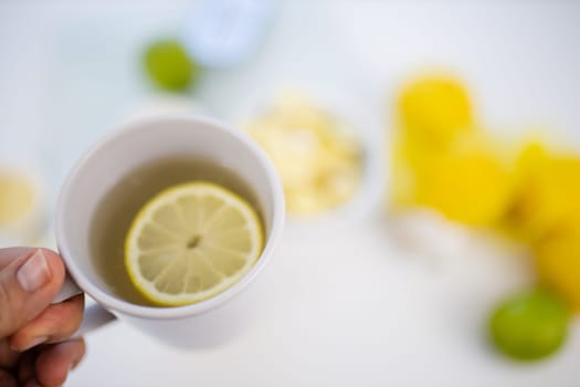 Female hand holding cup of lemon tea above blurry white table with limes and lemons. Woman holding white mug with lemon slice floating inside. Sweet citric beverages
