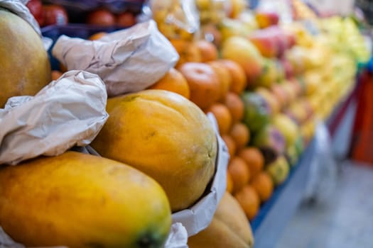 Colorful fruit stand with fresh papayas, tangerines, apples, mangoes, and more. Close-up of juicy-looking fruit for sale on stall inside classic Mexican market with blurry background. Healthy food