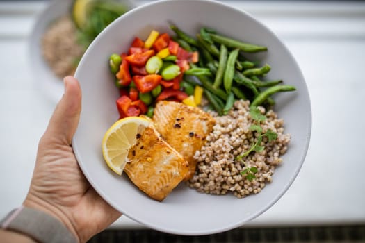 Hand holding salmon and buckwheat dish with green beans, broad beans, and tomato slices. Nutritious dish with vegetables and fish on white plate. Healthy balanced diet