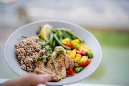 Hand holding chicken and buckwheat dish with green beans, broad beans, tomato, and pepper slices. Nutritious dish with vegetables and meat on white plate. Healthy balanced diet