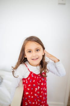 Cute little girl inside white room smiling and wearing red-flowered dress. Portrait of joyful young child in front of white wall. Happy kids at home