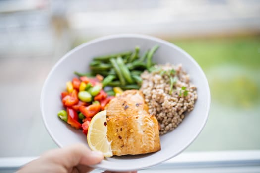 Hand holding salmon and buckwheat dish with green beans, broad beans, and tomato slices. Nutritious dish with vegetables and fish on white plate. Healthy balanced diet