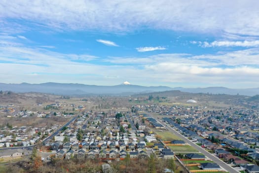 Peaceful small town under slightly cloudy sky and with mountains as background. Aerial view of slightly cloudy blue skyline above the suburbs. Rural town landscape