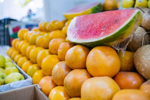 Close-up of colorful stand of fresh apples and oranges with sliced watermelons on top. Juicy-looking fruit for sale on stall with blurry background inside classic Mexican market. Healthy food