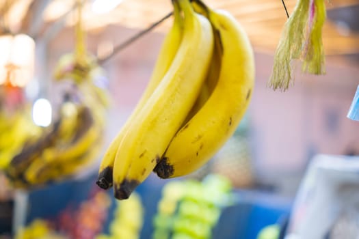Bunch of bananas hanging from thin rope with more blurry fruit as background. Fresh yellow fruit for sale inside classic Mexican market. Healthy food